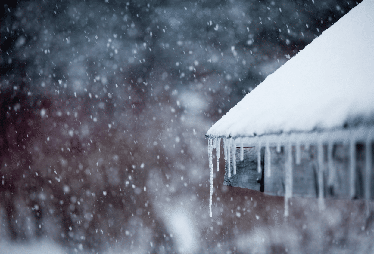 Snow-covered roof with long icicles forming along the edge during a winter snowfall.